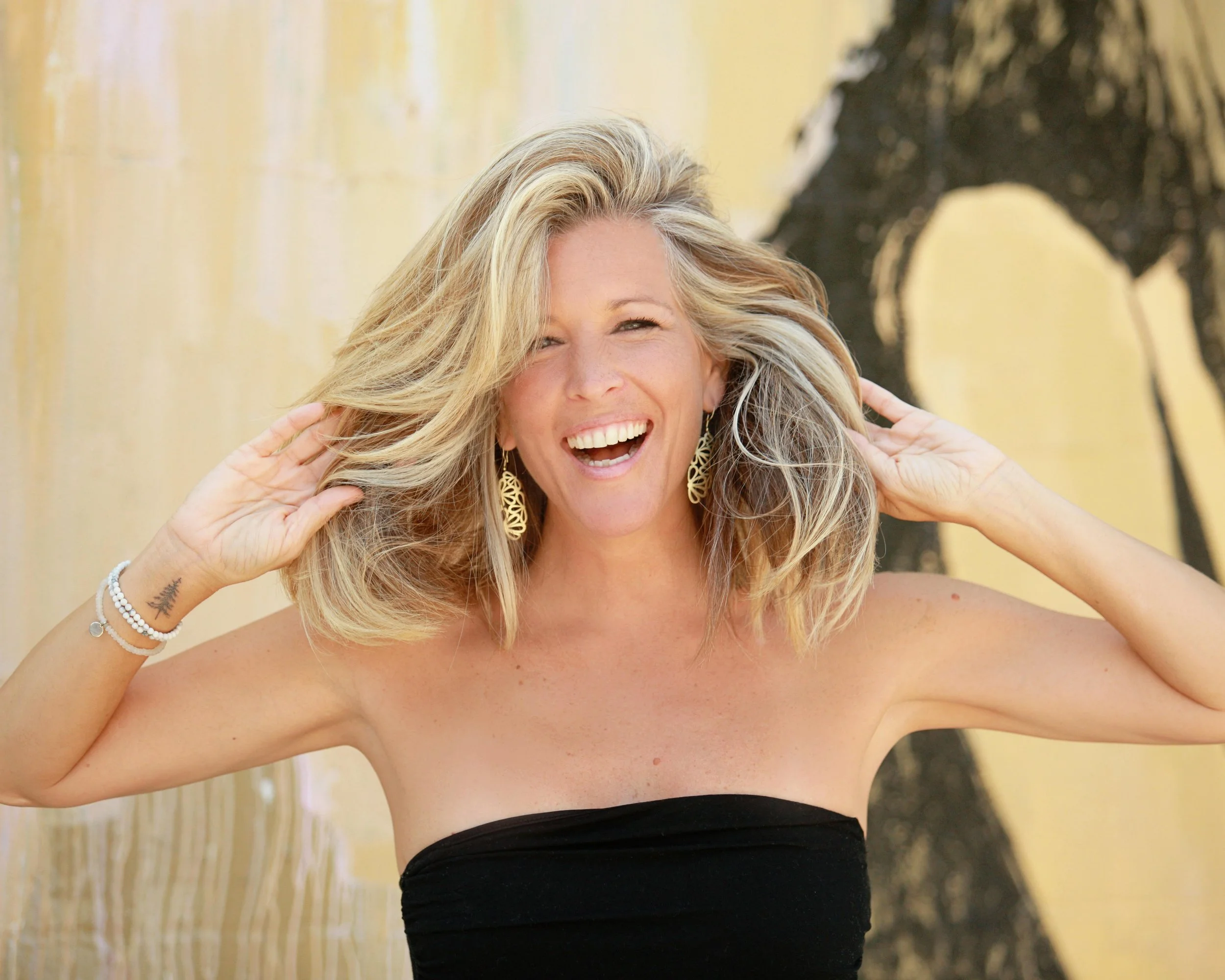 Woman with blonde hair, smiling, wearing gold earrings, in a black strapless top, standing outdoors near a tree.