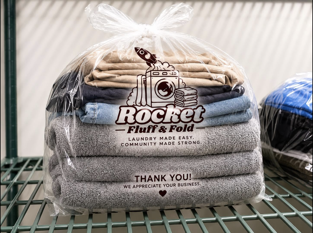 Stacked folded towels in different colors inside a clear plastic bag with a Rocket Fluff & Fold laundry service logo, placed on a metal shelf.