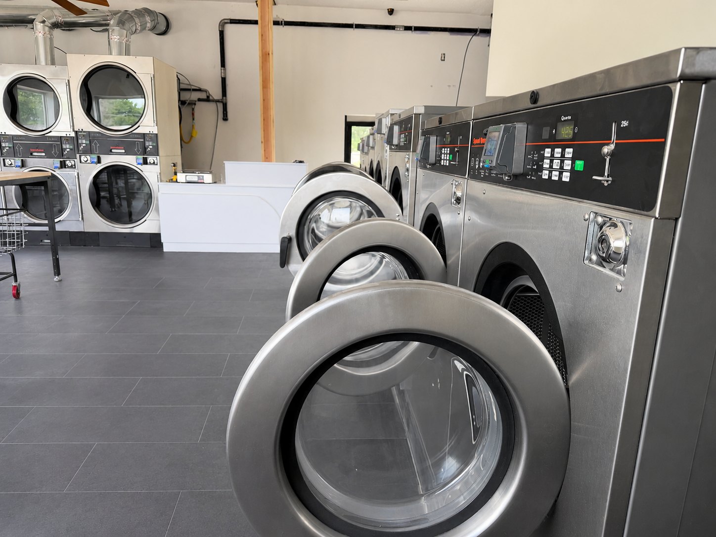 Multiple industrial washing machines and dryers in a laundromat with gray tiled floor, including front-loading washers and top-loading dryers, some with digital displays and control panels.