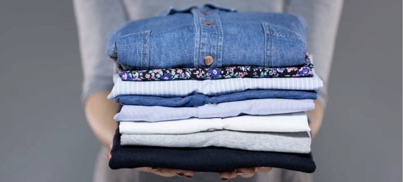 Person holding a neatly folded stack of various clothing items, including shirts and sweaters in different colors and patterns.