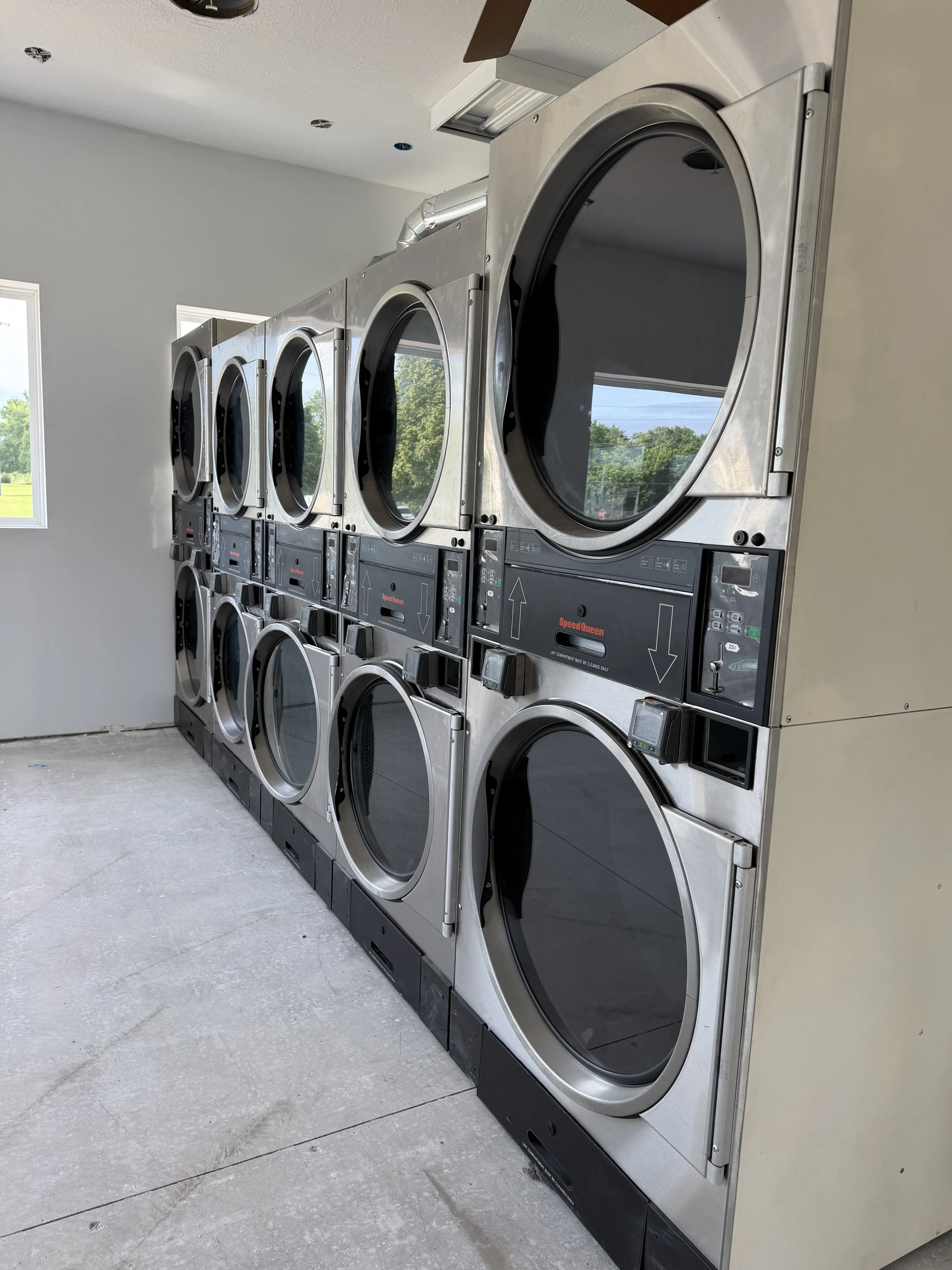 A row of front-loading commercial washing machines in a laundry room, with a window showing trees outside.