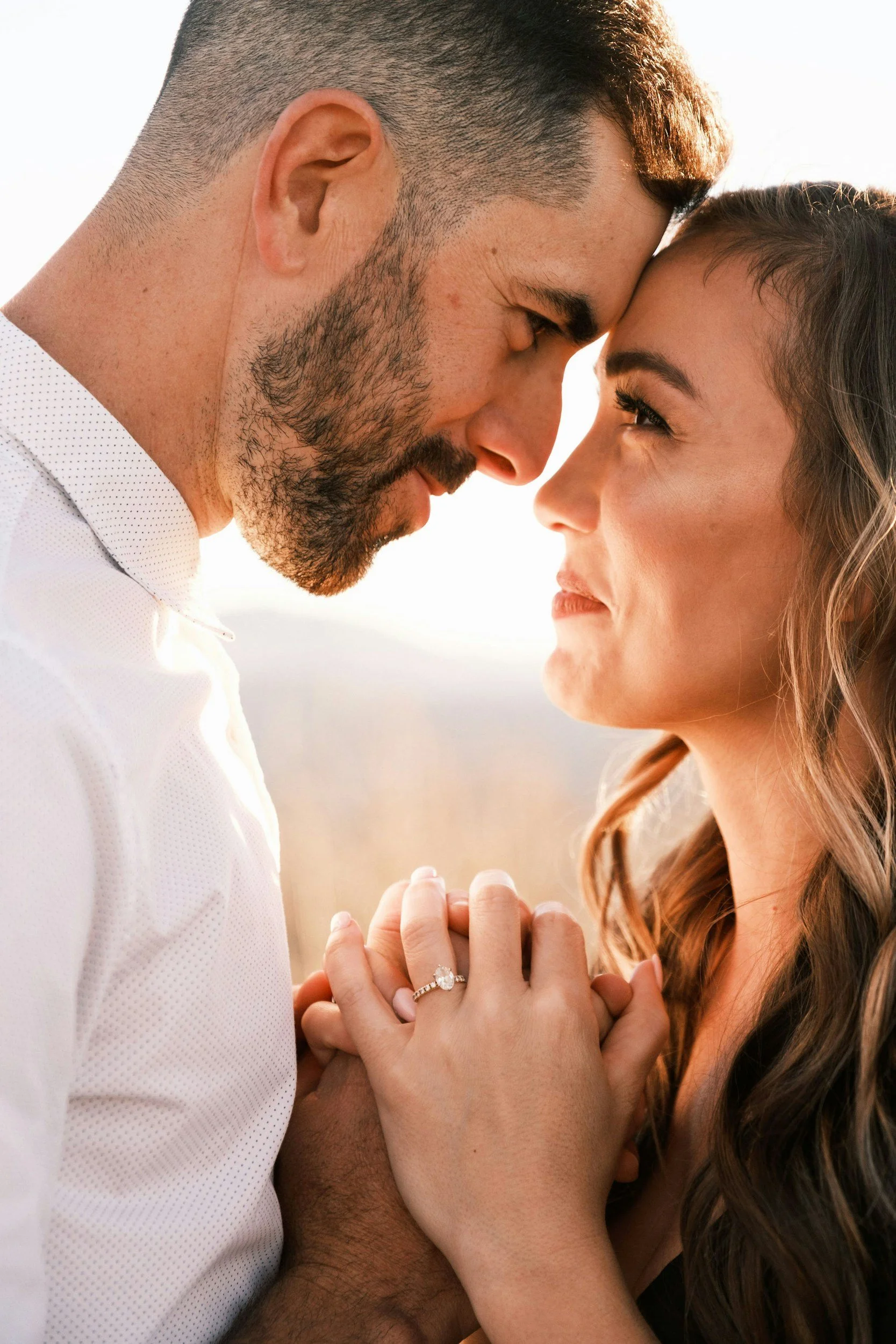 A couple with their foreheads touching, holding hands, with a bright background, and the woman showing an engagement ring.