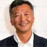 A smiling man with short, gray hair, wearing a suit and white shirt, in front of a whiteboard.