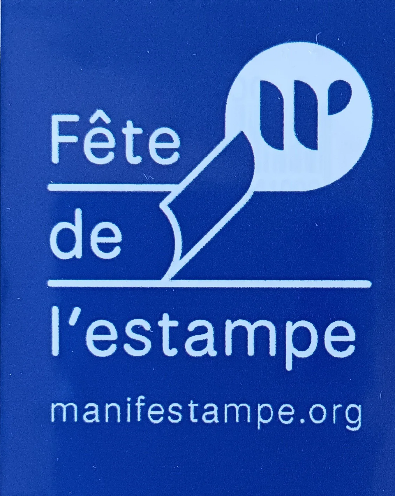 Ein blauer Hintergrund mit weißem Text und einem Logo, das eine Hand zeigt, die einen Stempel hält. Der Text lautet: "Fête de l'ouvrage" und darunter die Website-Adresse "manifestampe.org".