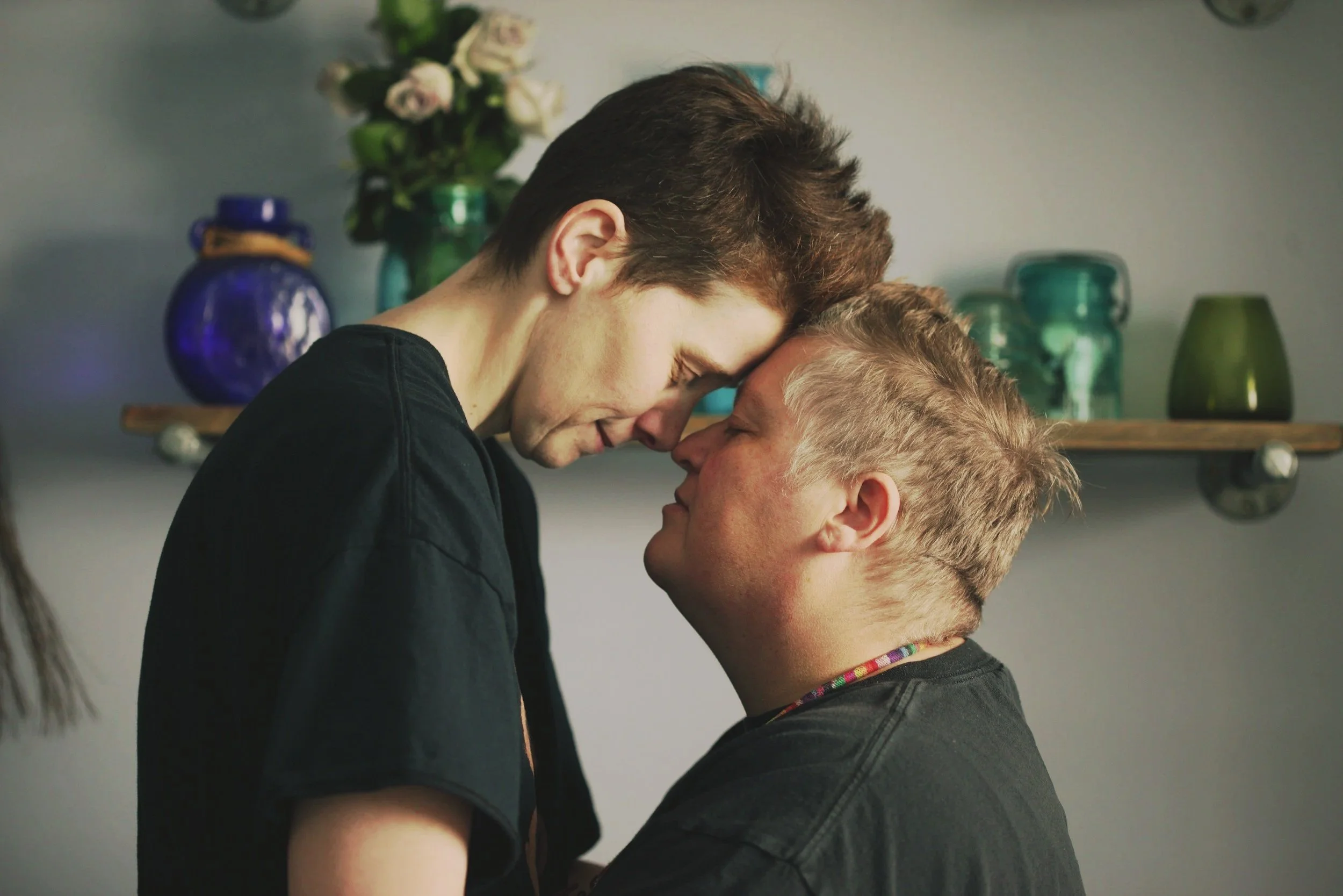 A couple are touching foreheads while facing each other, with closed eyes and gentle expressions. They are indoors, with a shelf holding vases and flowers in the background.