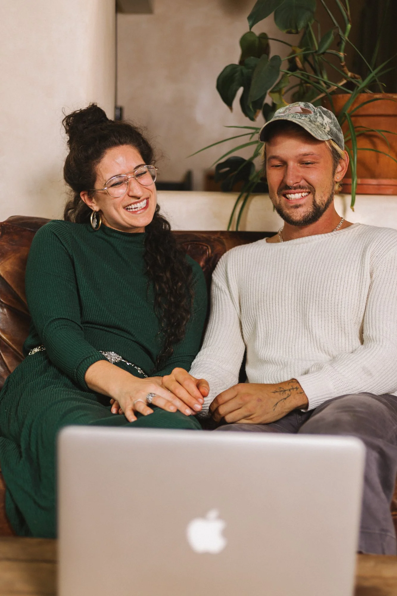 A couple sitting on a couch, holding hands, smiling and looking at a laptop screen.