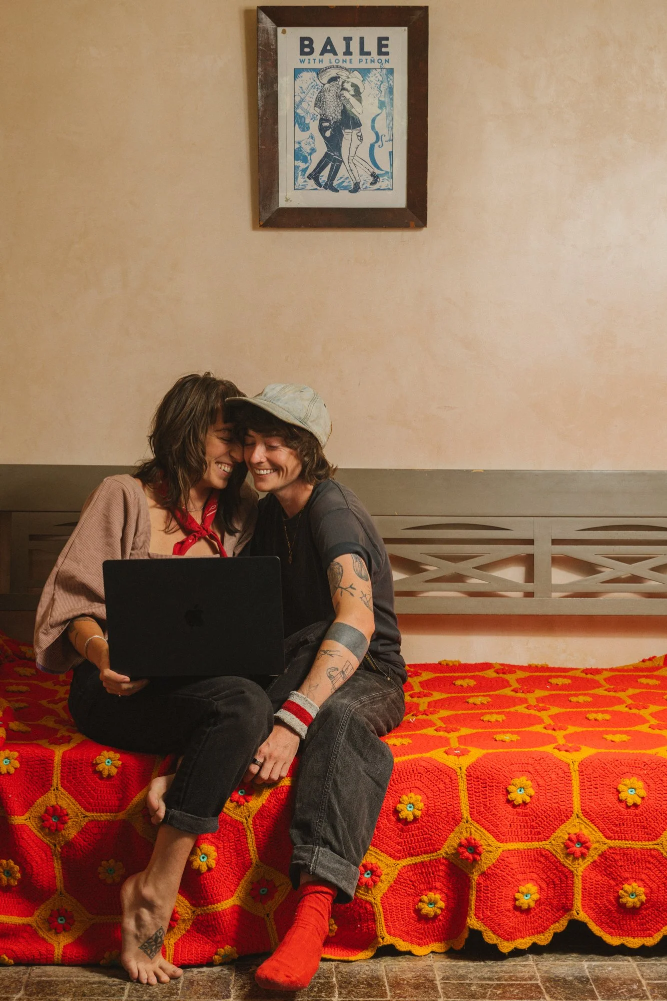 Two sitting on a bed, smiling and leaning close together, looking at a laptop. The bed has a red and orange crocheted blanket with yellow and blue flower details. One woman wears a gray hat, a black T-shirt, and has tattoos on her arm, while the other wears a pinkish blouse and a red bandana. Behind them is a beige wall with a picture frame hanging, containing a poster titled 'Baile with Lone Pinon'.