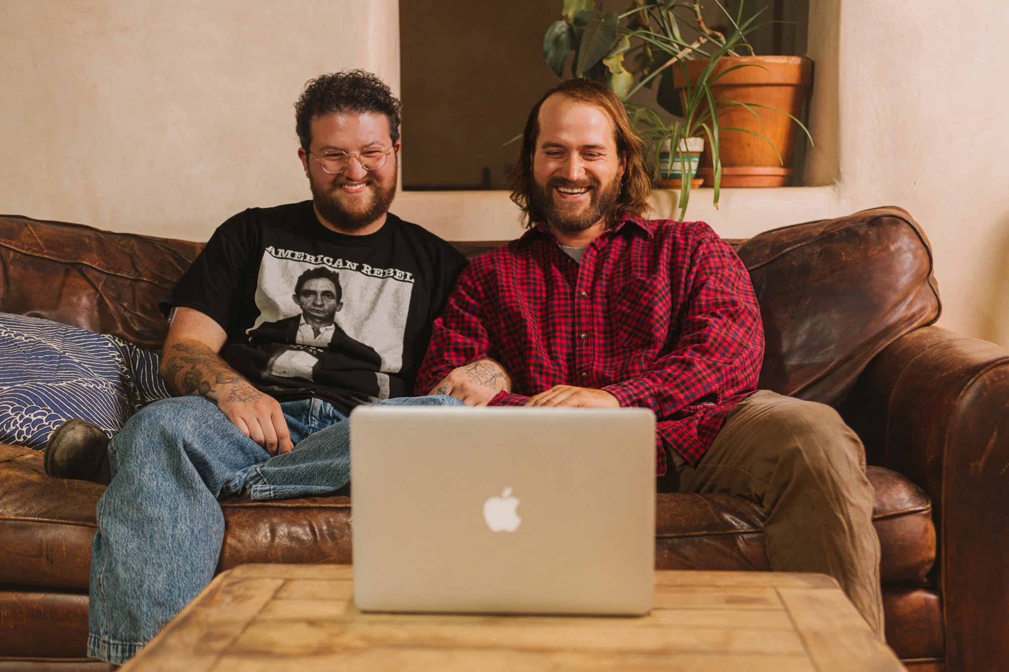 Two people sitting on a couch, looking at a laptop with smiles. One man wears a black T-shirt with a photo of Abraham Lincoln and the words "American Rebel." The other man wears a red and black checkered shirt. A potted plant is in the background.