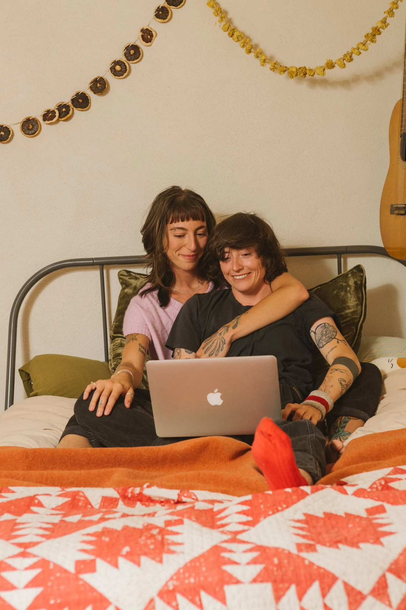Two queer women sitting on a bed, sharing a laptop, smiling and close together, in a cozy room with decorative wall hangings and an acoustic guitar.