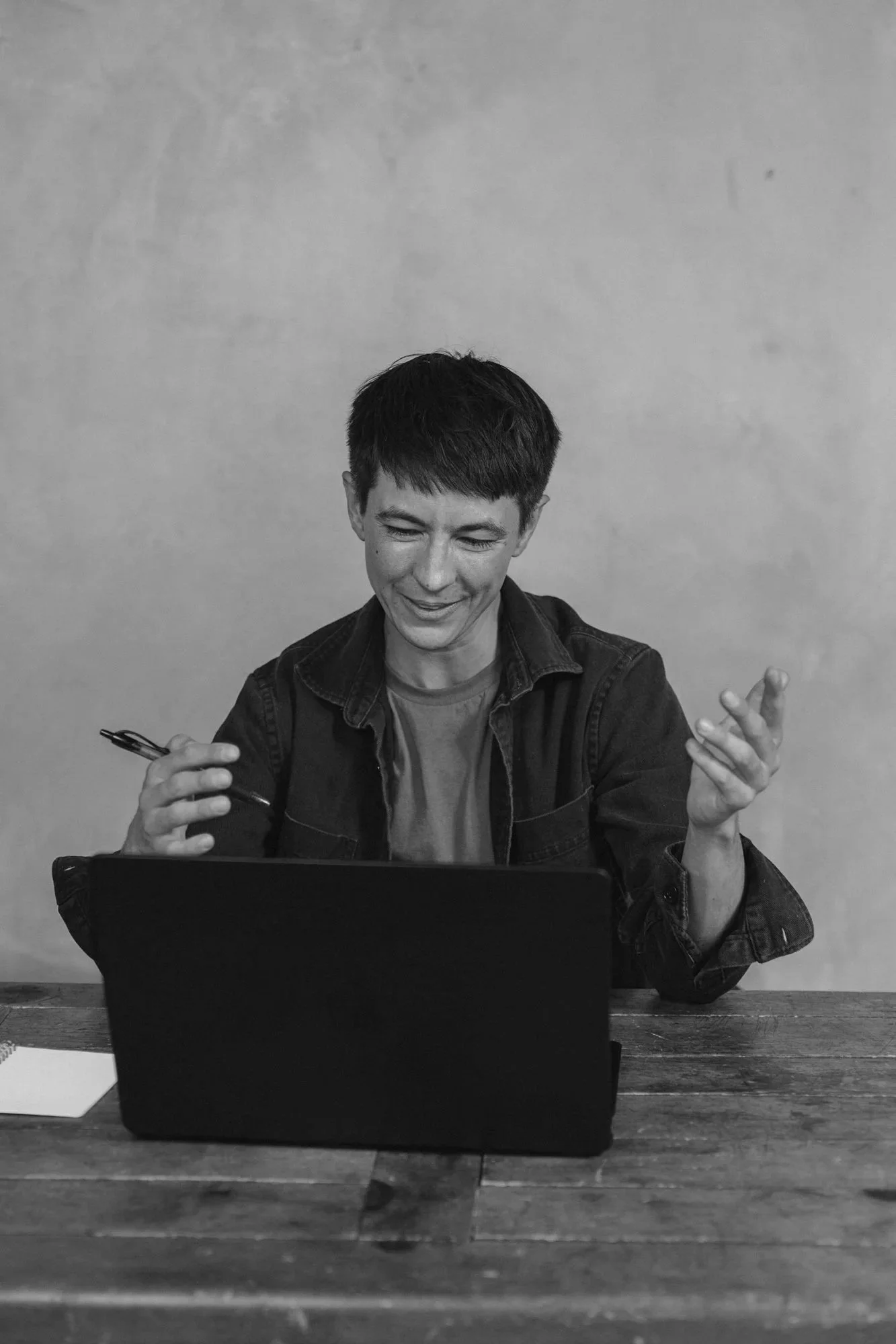 A person with short dark hair, smiling, sitting at a wooden table in front of a laptop, holding a pen in his right hand, with a notebook on his left, against a plain background.