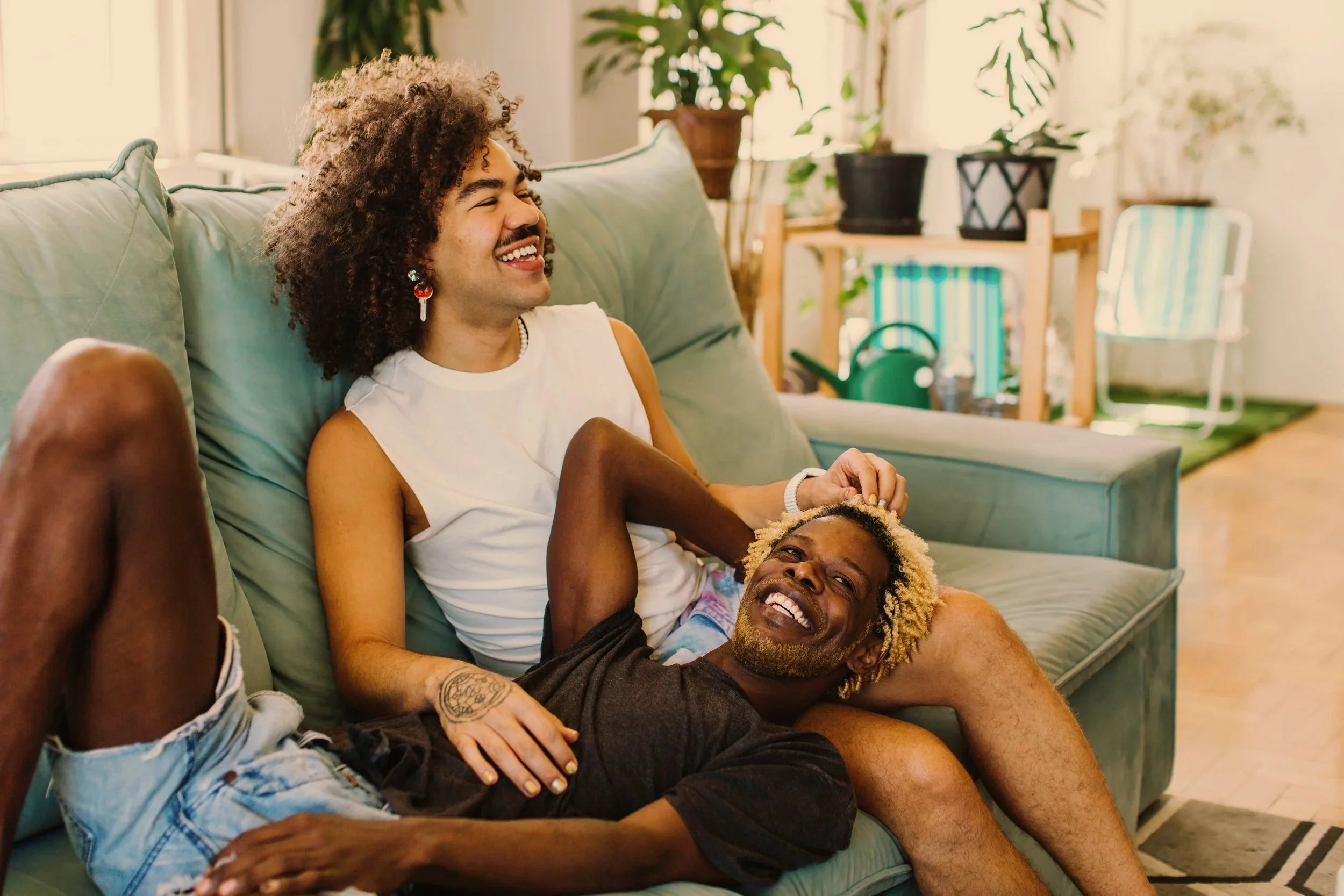 Two queer men with curly hair relaxing and laughing on a green sofa in a sunlit living room.