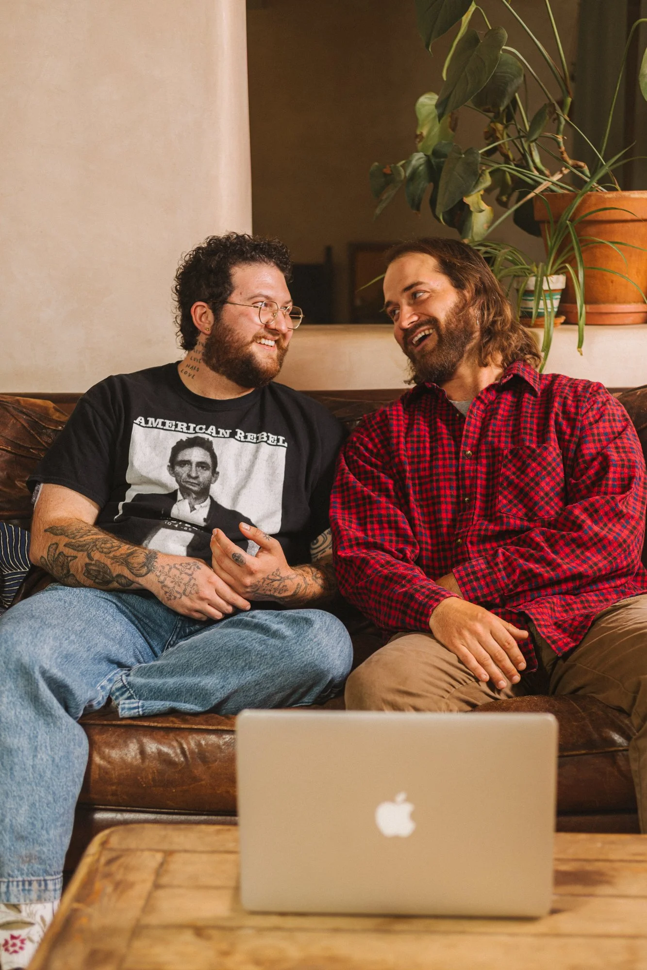 Two queer men sitting on a couch, smiling and talking, with a laptop in front of them, in a cozy room with plants.
