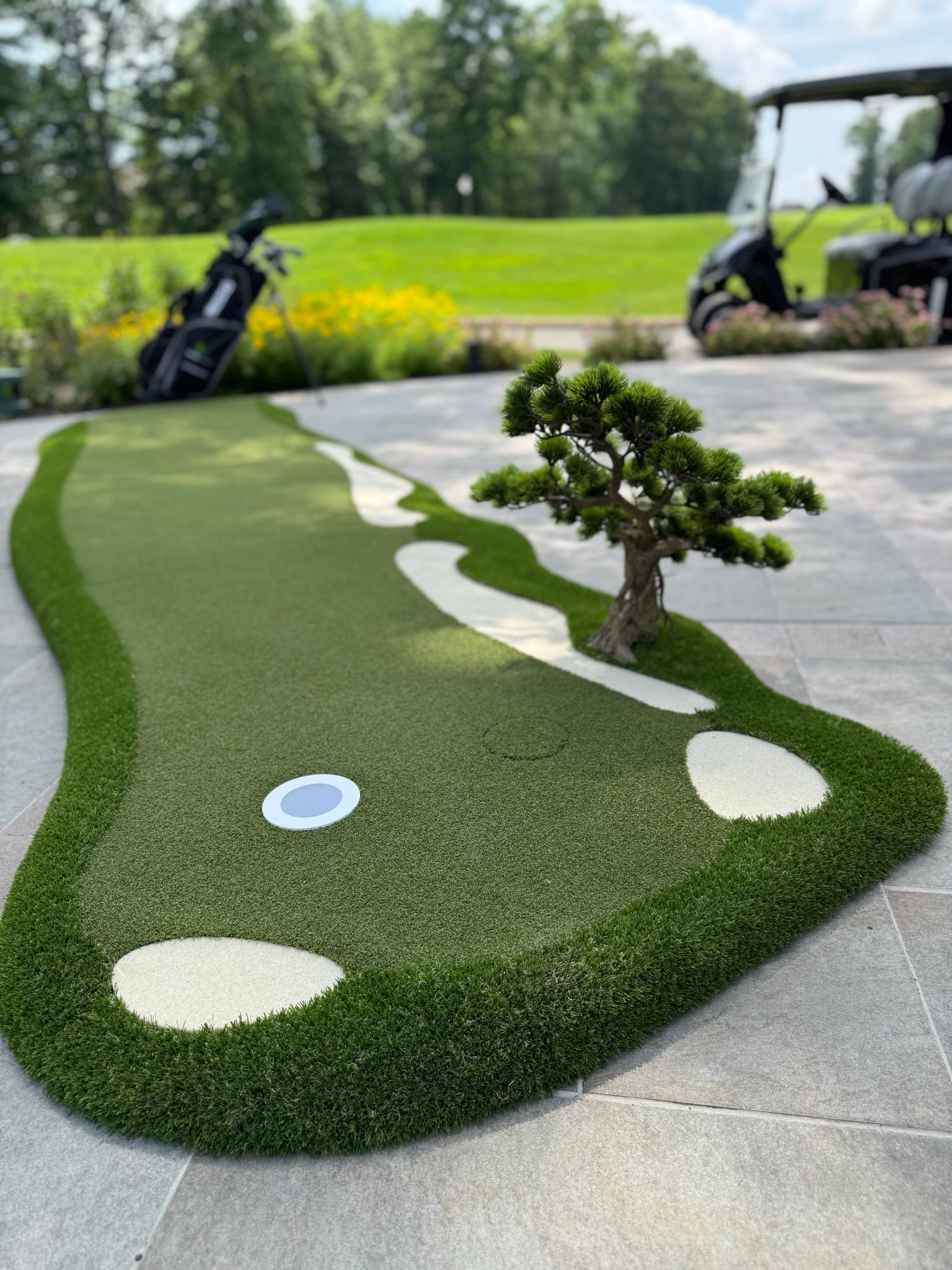 Artificial grass modular putting green, with a set of golf clubs behind it as well as a golf course and golf cart in the background.