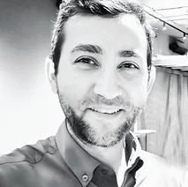 Black and white photo of a smiling man with short hair and a beard, wearing a collared shirt, indoors.