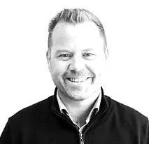 A smiling man with short hair wearing a collared shirt and a jacket, in black and white