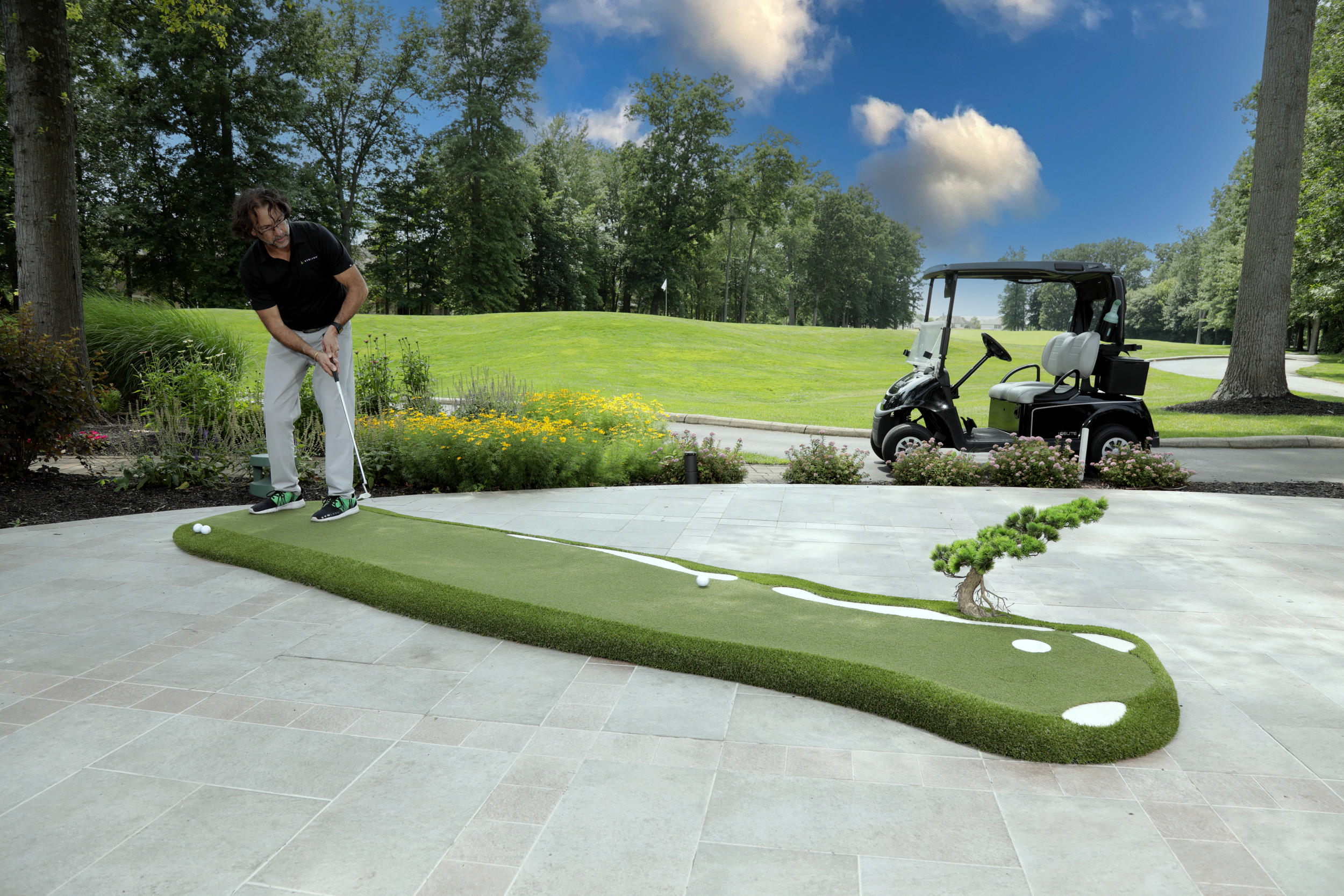 man putting with golf club on portable putting green ontop of a patio, golf cart, trees and golf course in background