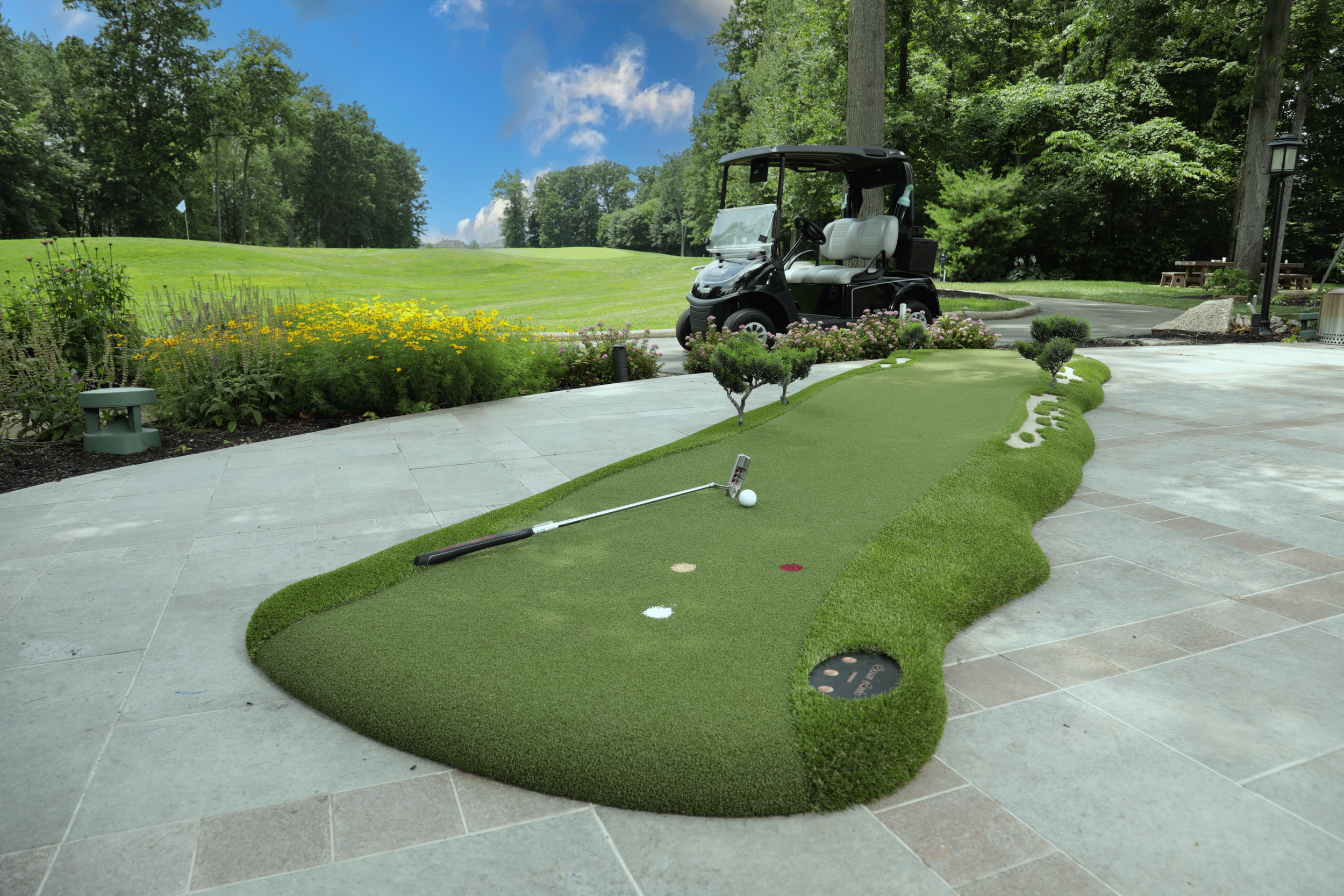 modular putting green on a patio with a golf course and golf cart in the background with a golf ball and golf club resting on top of the putting green