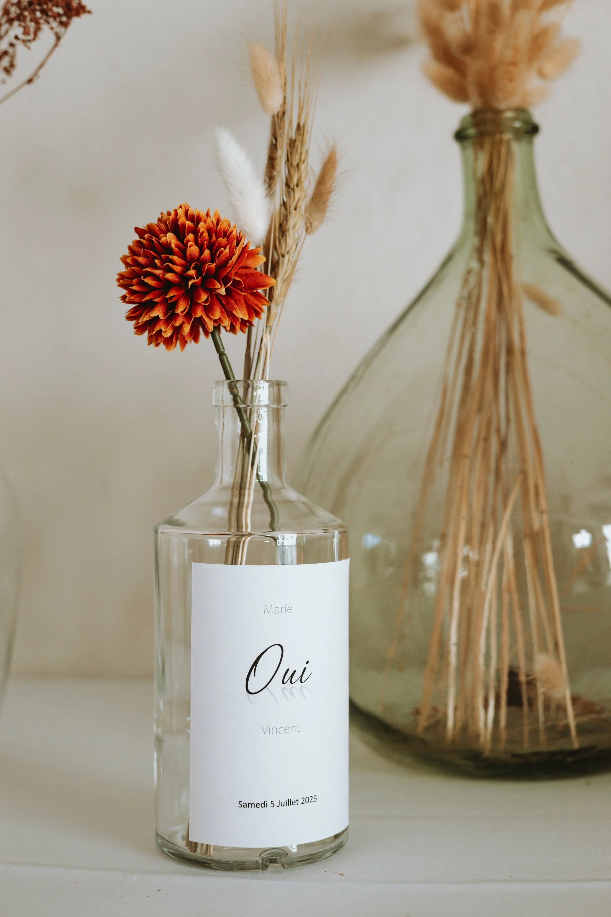 Vase en verre avec une étiquette indiquant 'Oui', contenant une seule fleur orange et quelques herbes sèches, à côté d'une autre grande bouteille en verre contenant des herbes sèches.