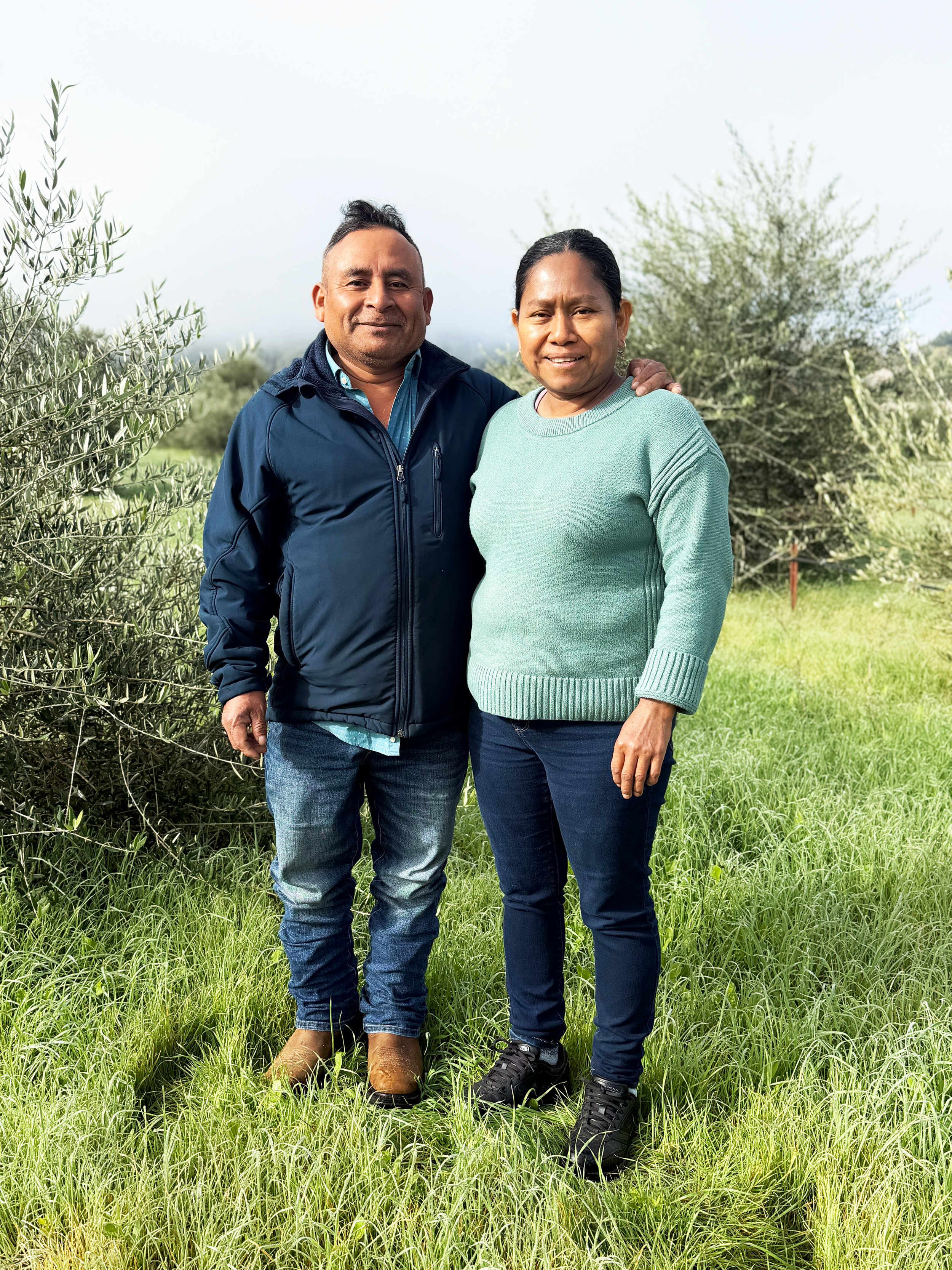 A man and woman standing outdoors on a grassy field with bushes and trees in the background, smiling at the camera.