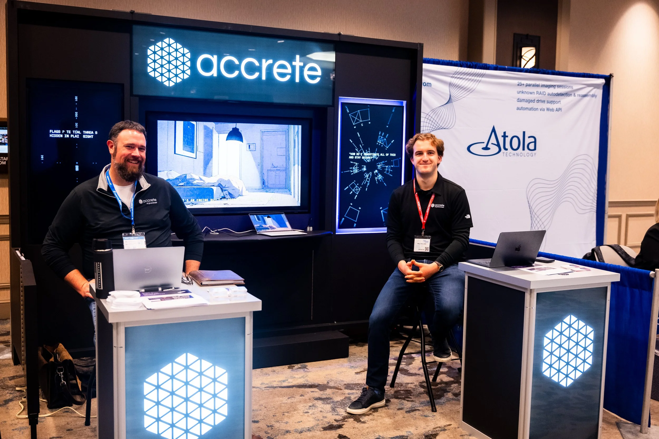 Two men wearing black shirts with Accrete logo sitting at tables with laptops at a trade show booth, with a large screen displaying digital images and the Accrete logo above.