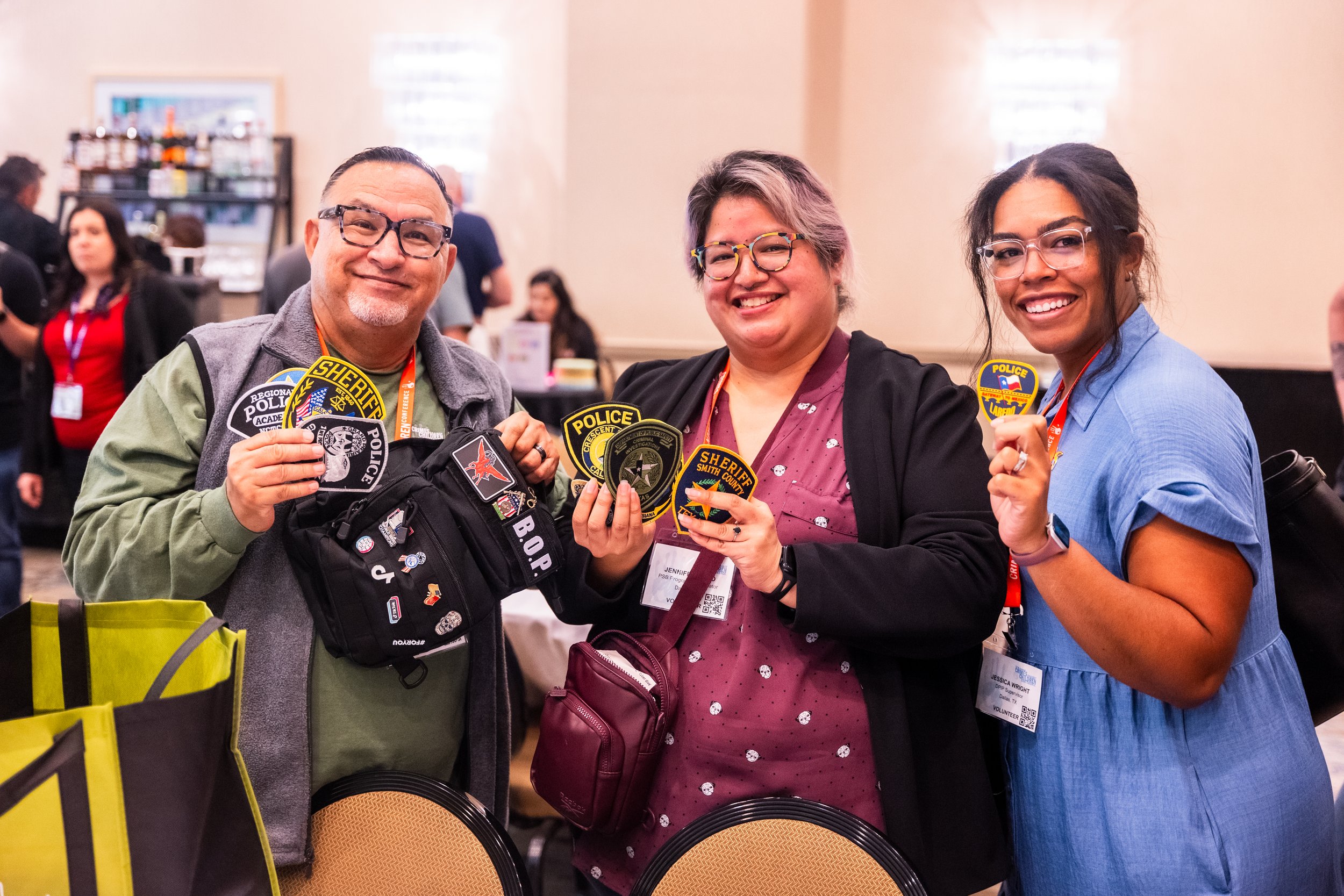 Three smiling people at a conference holding police patches, standing behind two chairs, with conference badges around their necks.