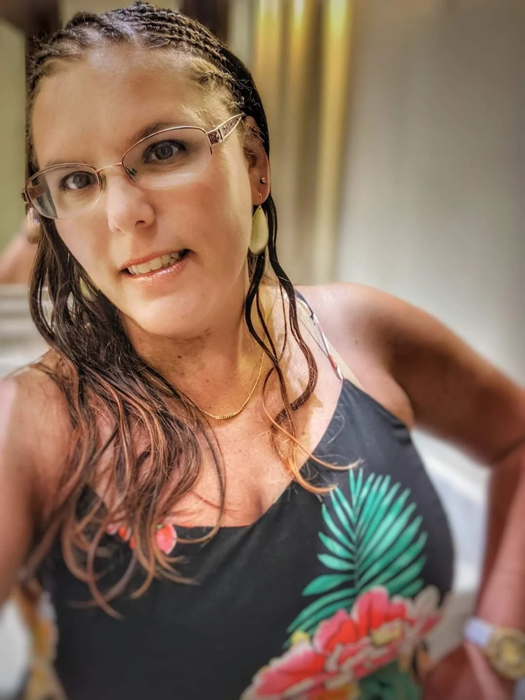 A woman taking a selfie, wearing glasses, earrings, a necklace, and a floral dress with pink and green designs, with a blurred background.