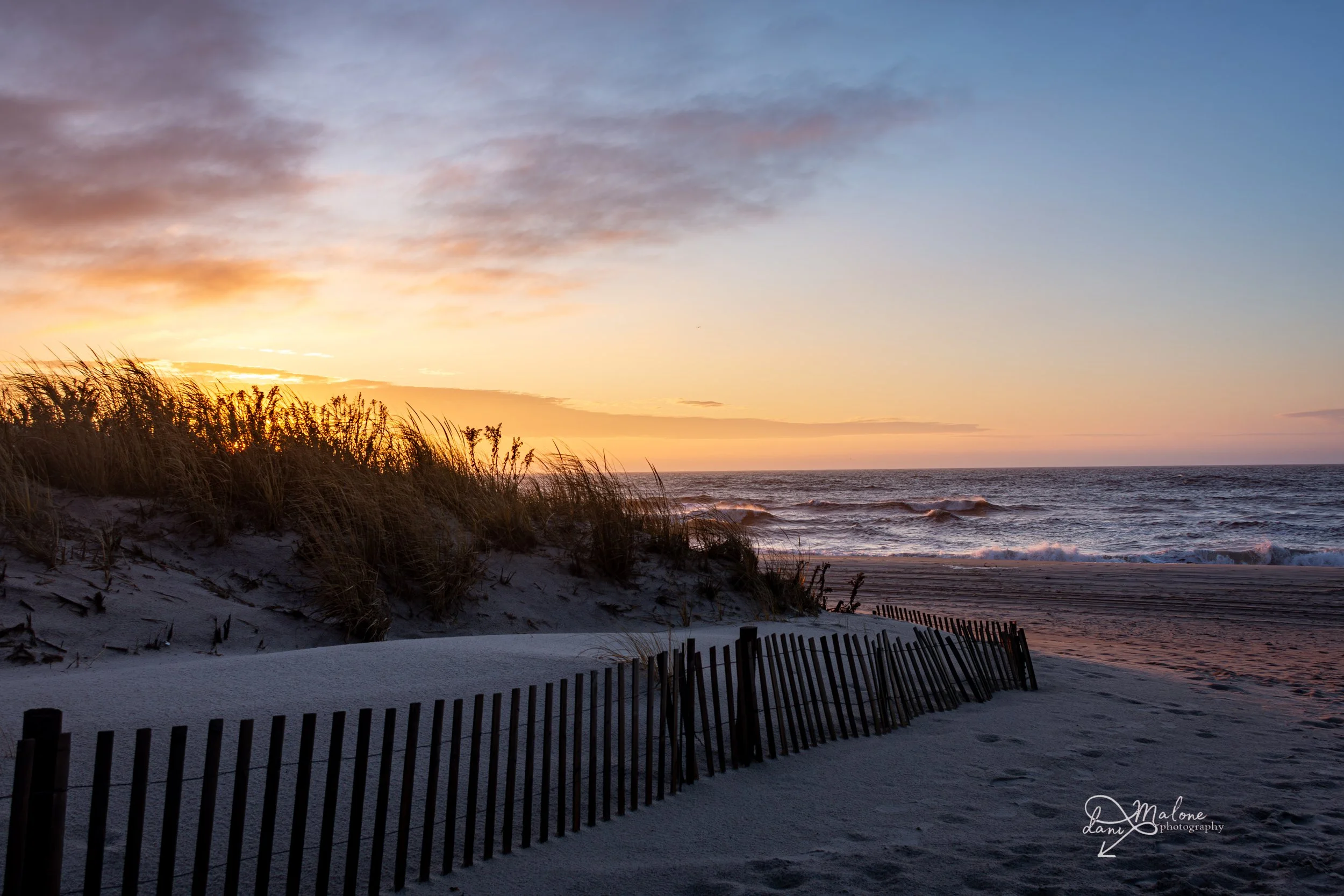 A sunset over the ocean with waves crashing and a sandy beach with a wooden fence and grassy dunes