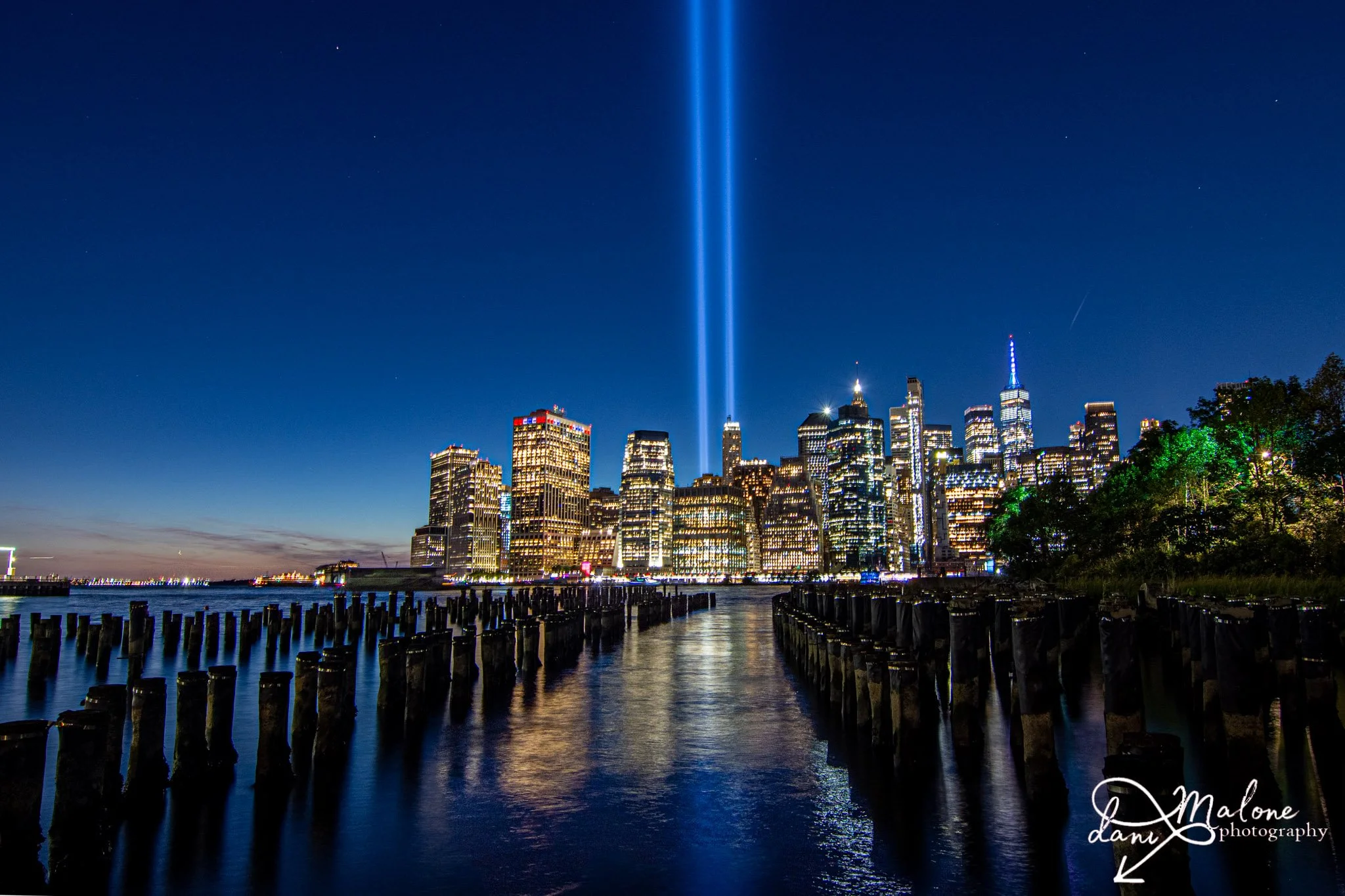 Nighttime view of Manhattan skyline with the World Trade Center's One World Trade Center illuminated, reflecting in the Hudson River, with the Manhattan Bridge in the background and beams of blue light shooting up into the sky.
