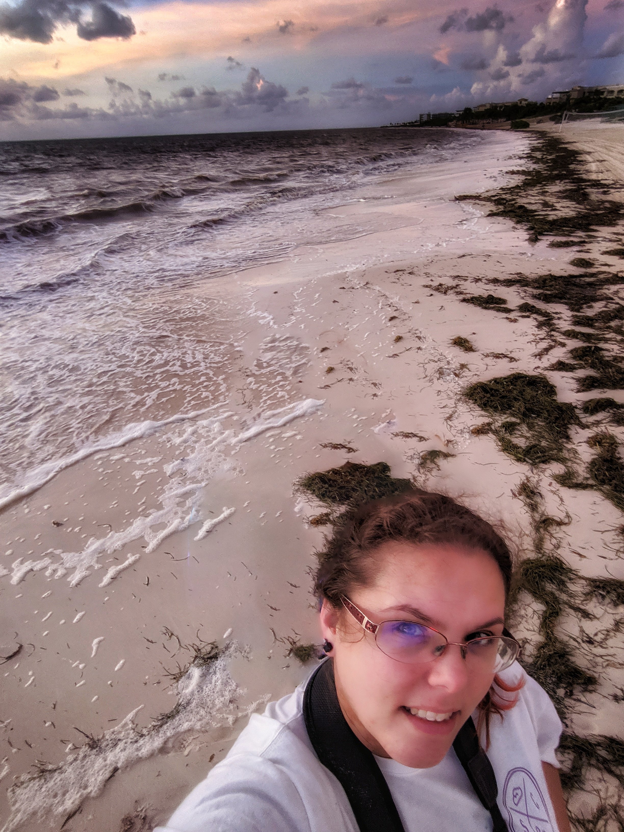 A woman taking a selfie on a sandy beach during sunset, with seaweed scattered along the shore and waves gently crashing, under a colorful cloudy sky.