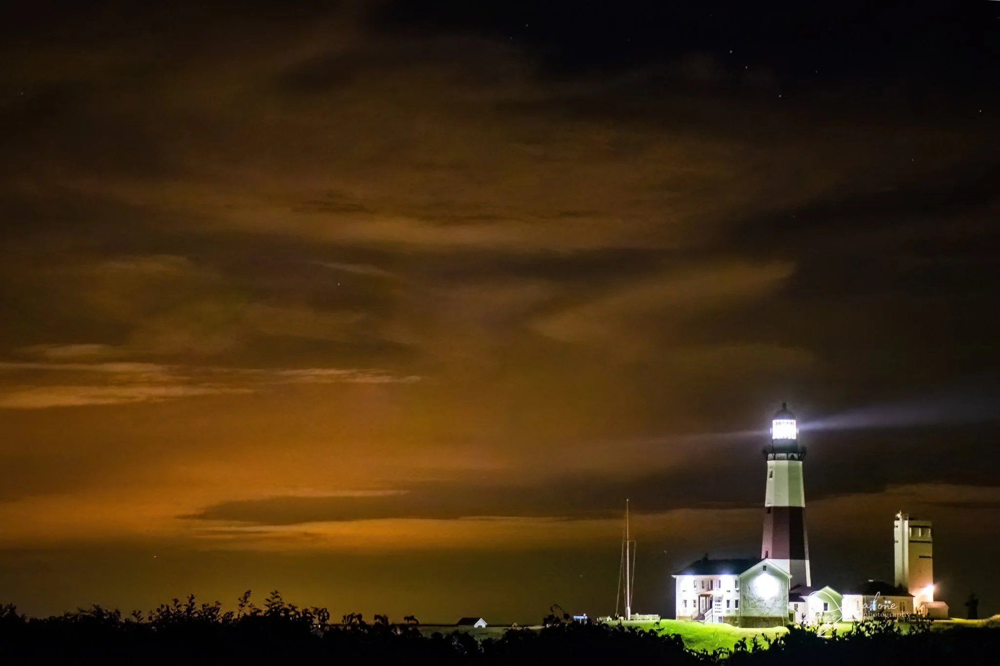 Nighttime view of a lighthouse with a bright light on top, attached to a white building, against a cloudy sky with some stars visible.