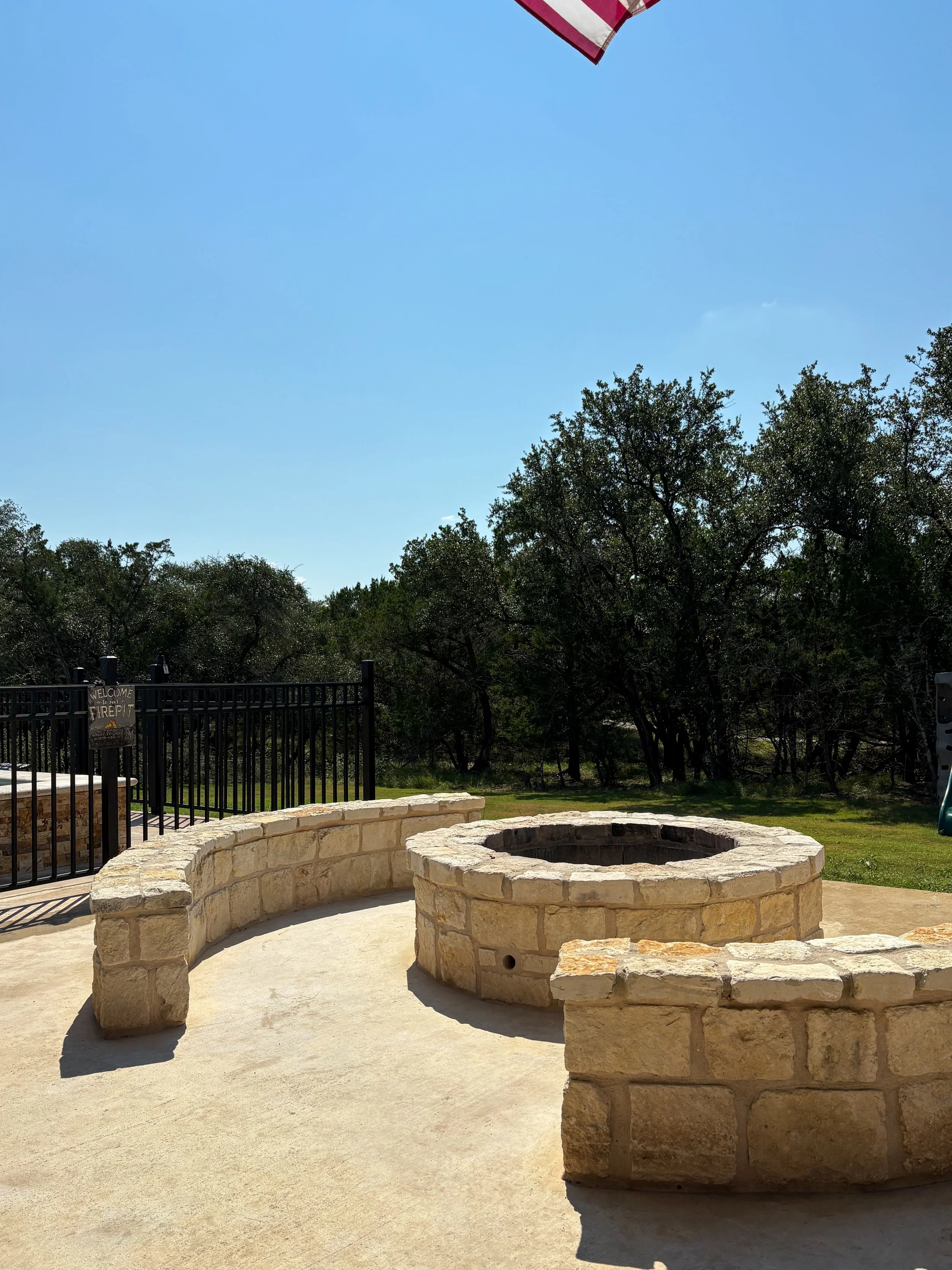 Stone fire pit with curved stone bench seating in outdoor patio area, trees and a blue sky in the background, and part of an American flag visible in the top right corner.