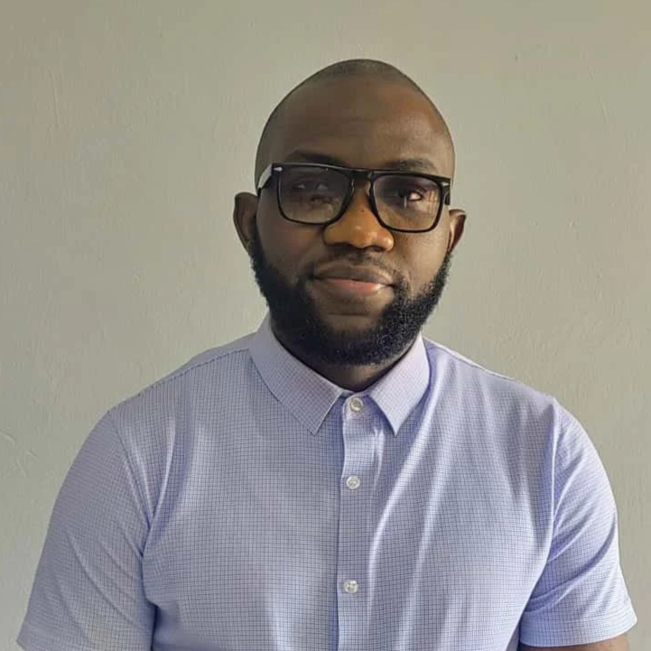 A man with a beard and glasses wearing a light purple button-up shirt, sitting against a plain wall.