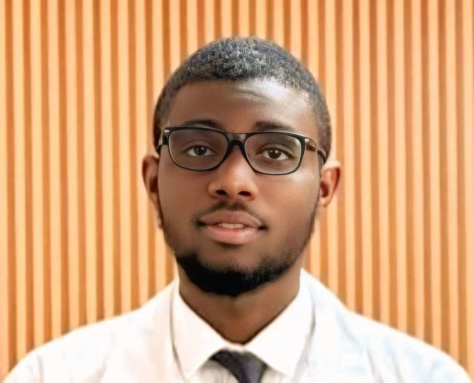 A young man with short hair, glasses, and a beard, wearing a white dress shirt and tie, standing in front of a wooden panel background.