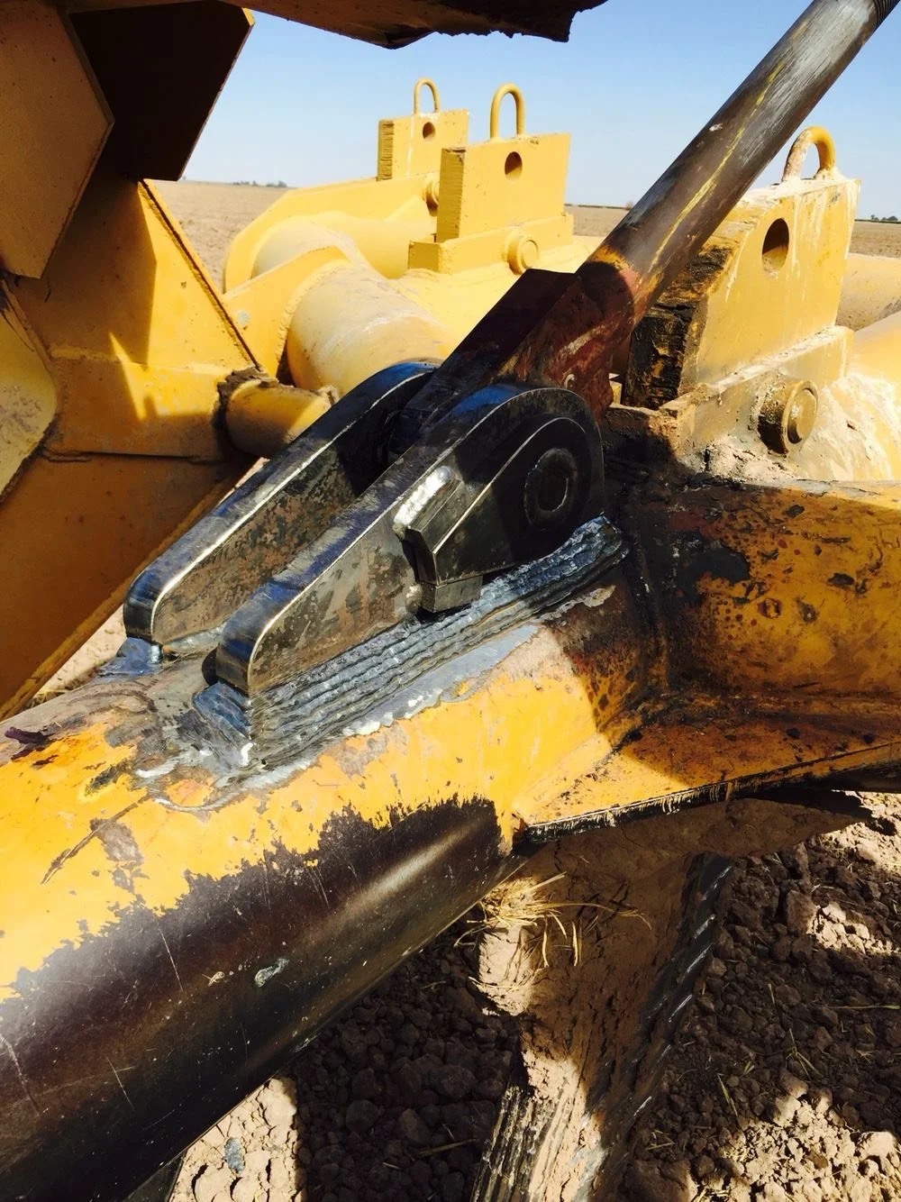 Close-up of a yellow construction or agricultural machine part showing hydraulic components and metal joints with some dirt and rust.