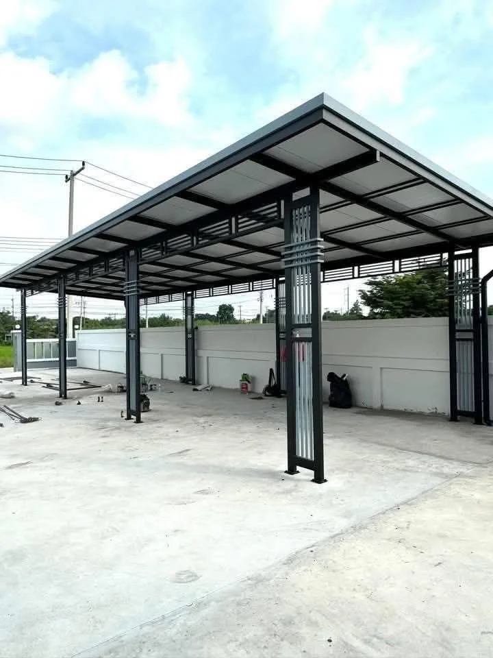 A metal carport structure with a roof, positioned on a concrete surface, with a white wall and open outdoor area in the background.