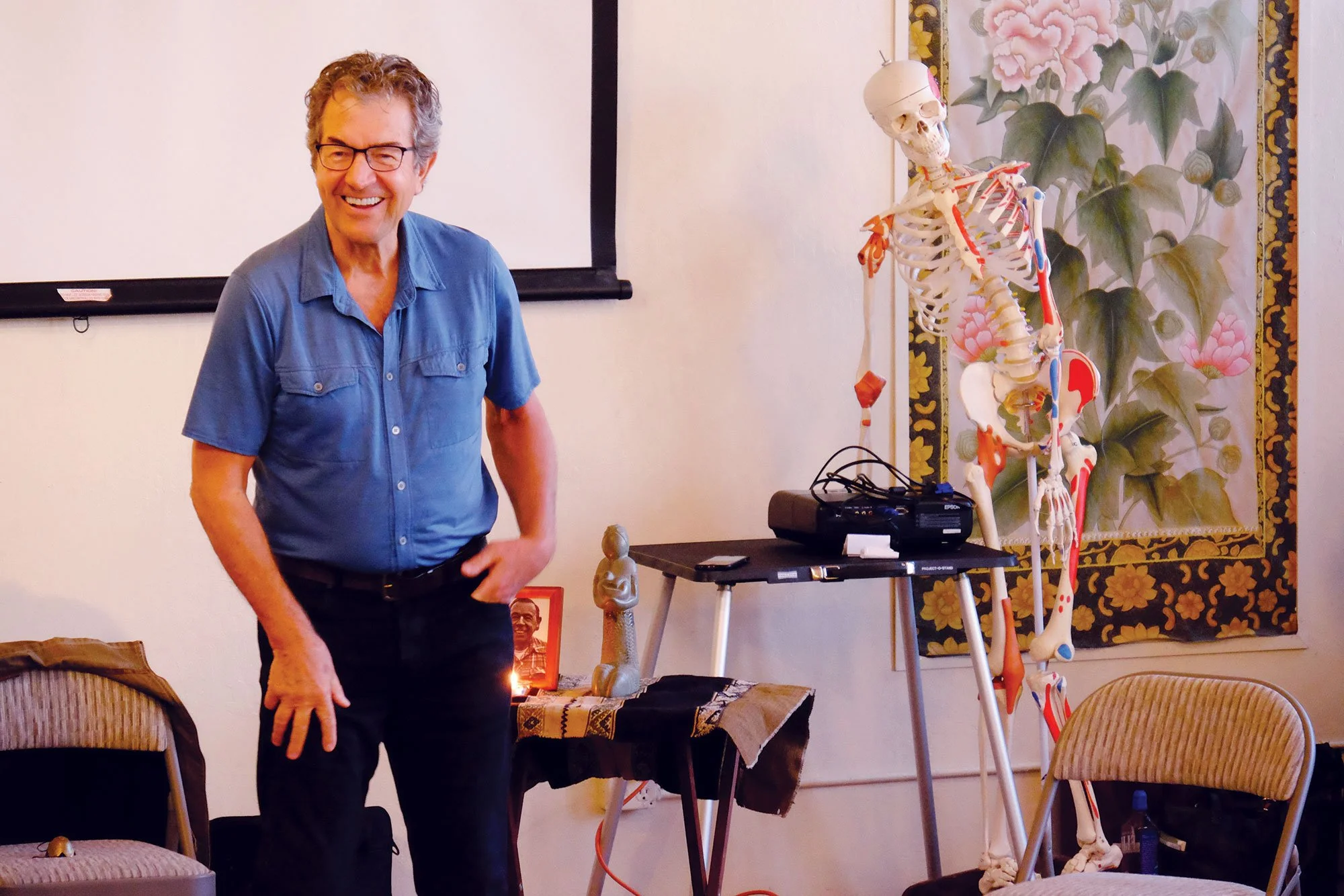 A man wearing glasses and a blue shirt standing next to a table with a lit candle, in a room with a skeleton model and floral wallpaper in the background.