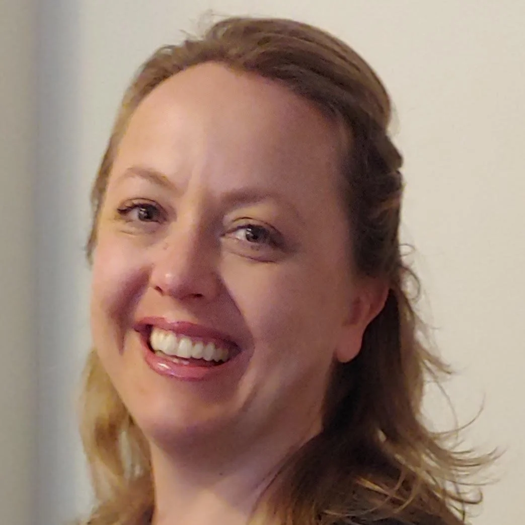 Close-up of a smiling woman with light brown hair, showing her teeth, against a neutral background.