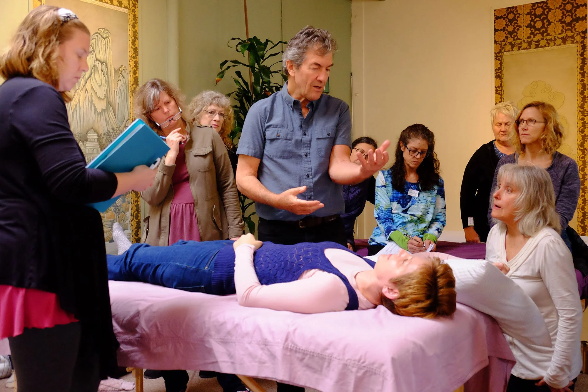 A group of people gathered around a woman lying on a massage table, engaging in what appears to be a group workshop or demonstration, with some participants taking notes.