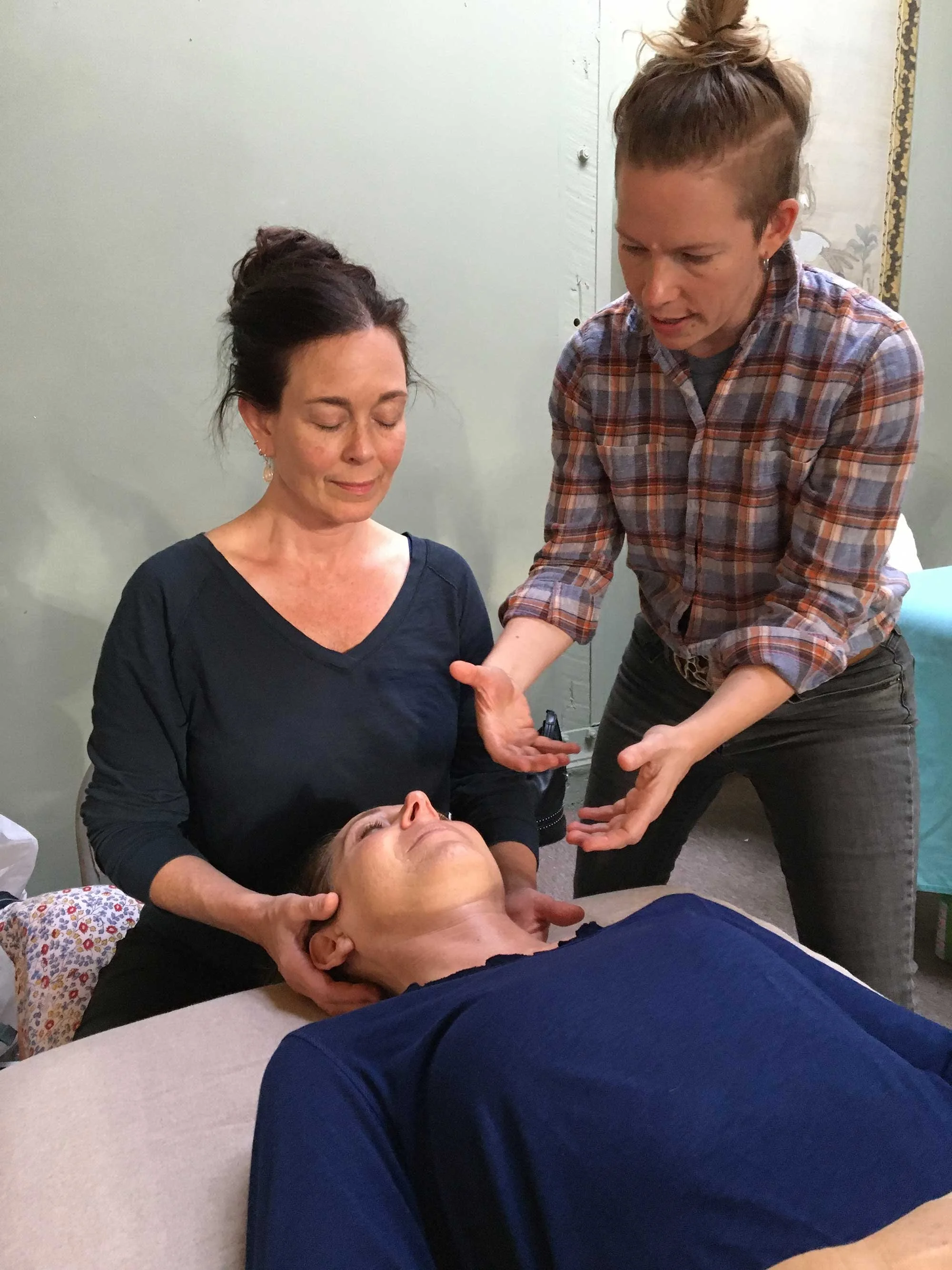A woman with dark hair in an updo and earrings is receiving a neck massage from a person in a plaid shirt, while another person with long hair lying down with eyes closed rests on a table.