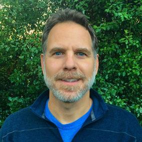 A middle-aged man with a beard and short hair, smiling outdoors in front of green foliage, wearing a dark jacket over a blue shirt.