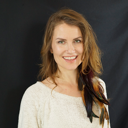 A woman with shoulder-length wavy hair and light skin smiling at the camera, wearing a cream sweater and a feather accessory on her shoulder, against a dark background.