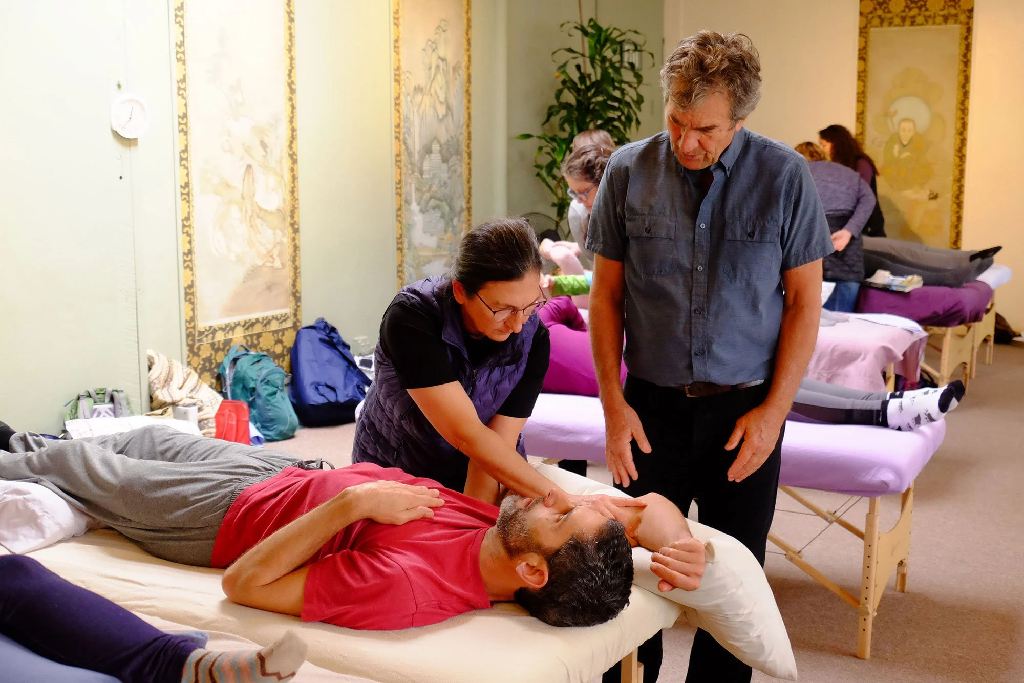 People participating in a massage or therapy session in a room with multiple beds, some people lying down and others standing or sitting nearby, with shelves and paintings on the walls.