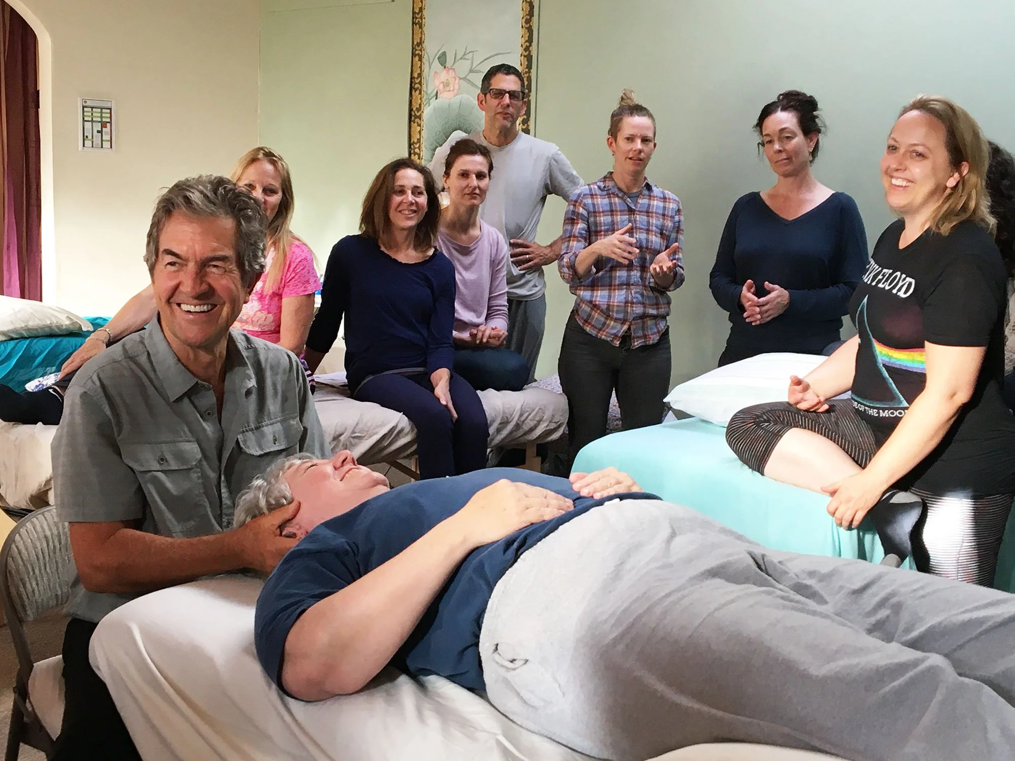 A group of people gathered around an elderly man lying on a hospital bed. They appear to be having a conversation, with some standing and some sitting. The mood seems warm and friendly.