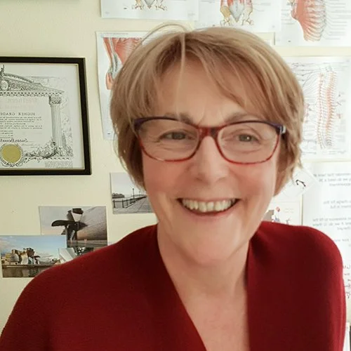 Smiling middle-aged woman with short blonde hair and red glasses, wearing a maroon top, in front of a wall with framed certificates and anatomical charts.