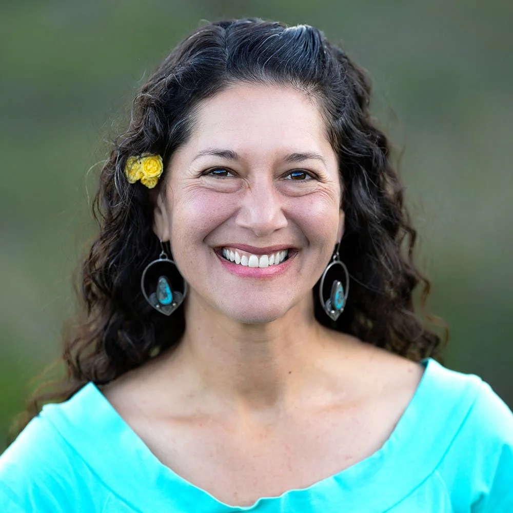 A woman with dark curly hair, wearing a blue top, yellow flower hair accessory, and turquoise earrings, smiling outdoors.