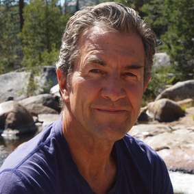 A middle-aged man with gray hair and a slight smile, outside near rocks and trees.