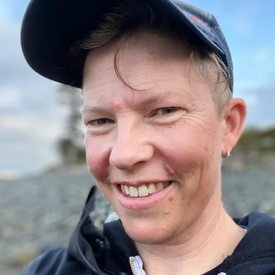 Close-up of a smiling person wearing a baseball cap outdoors with a cloudy sky in the background.