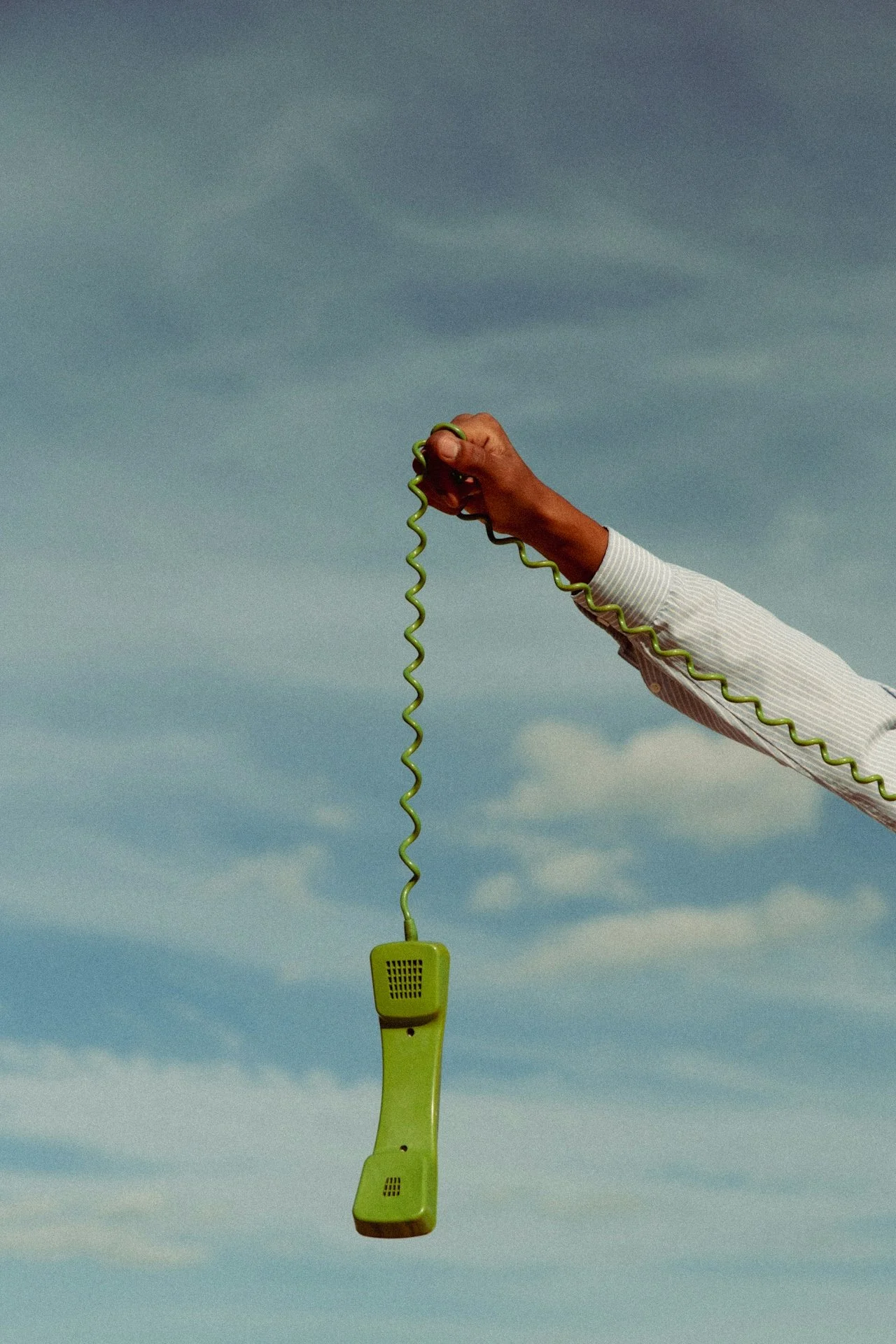 A person holding a vintage green corded telephone by the receiver against a partly cloudy sky.