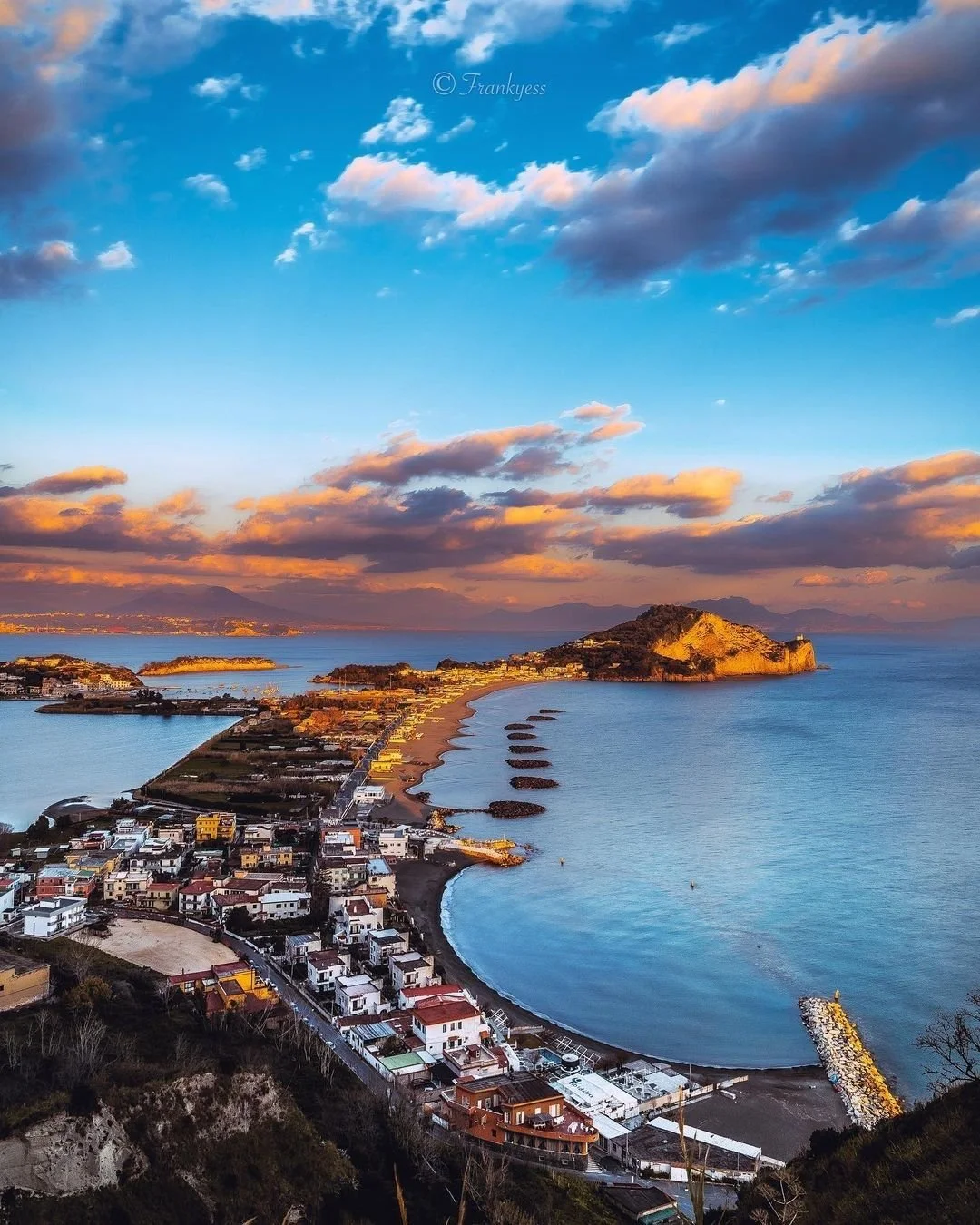 A coastal town with white buildings along the shoreline, showing a long sandy beach curve and a calm ocean, with an island and mountainous landforms in the background during sunset weather.