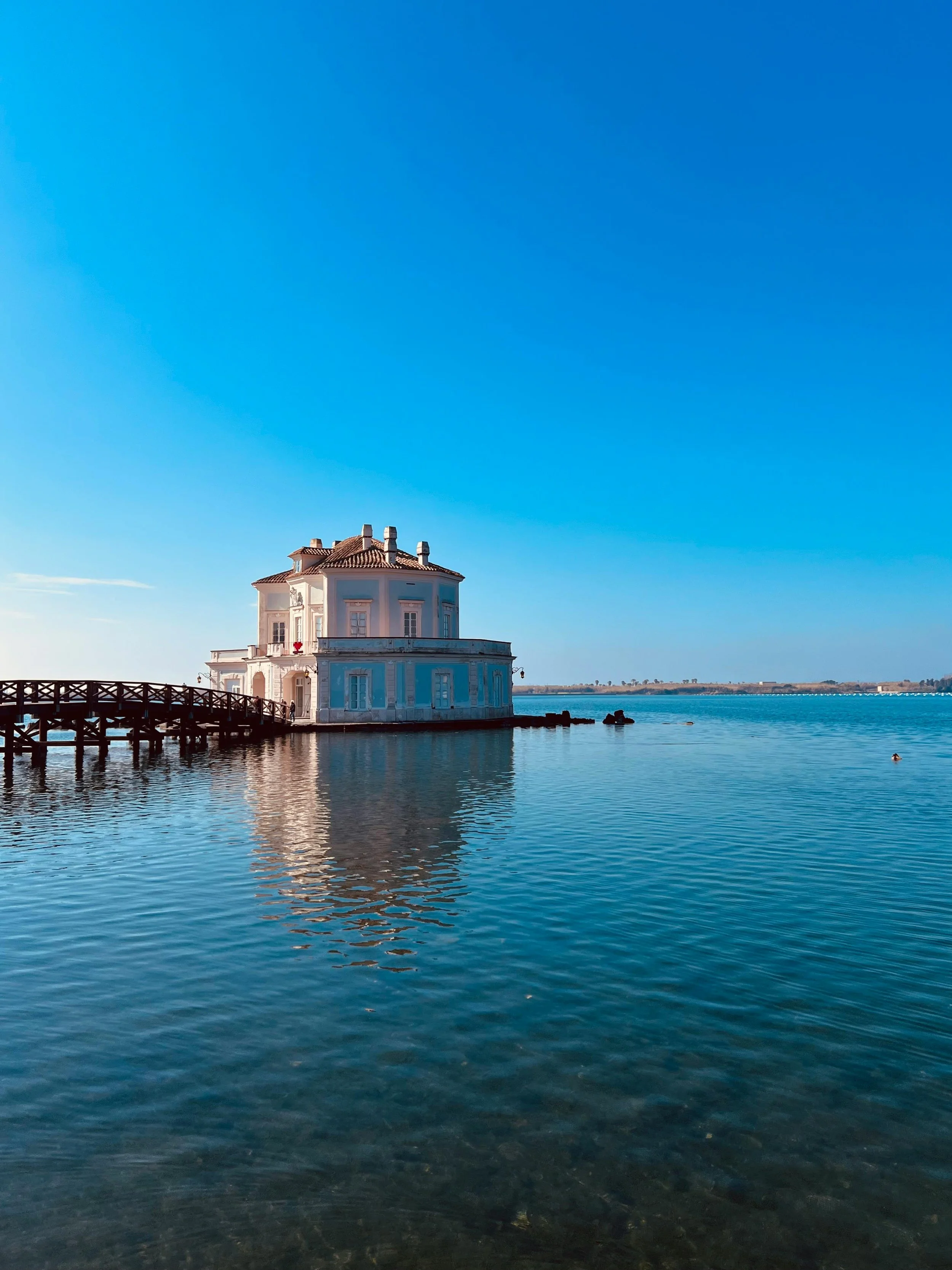 A white, multi-story building with a tiled roof, set against a large body of water, under a bright blue sky.